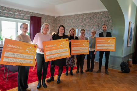 Sju personer står inomhus och håller stora orangea checkar med text på svenska.