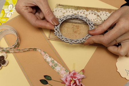 Två händer dekorerar ett kort med spets, ram och blommor vid ett pysselbord.