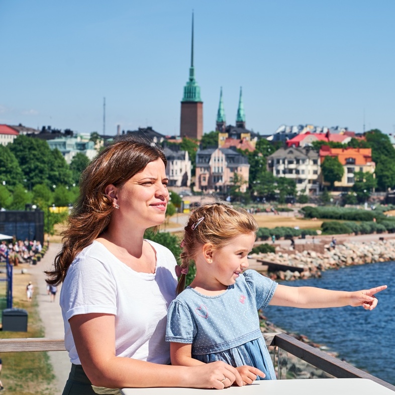 En kvinna och ett barn står vid vattnet och tittar ut över en stad i solsken.