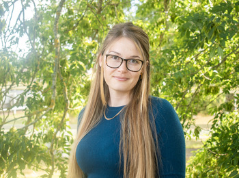 Kvinna med långt blont hår och glasögon står utomhus framför gröna träd, bär blå tröja och halskedja.