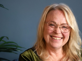Leende äldre kvinna med långt blont hår och glasögon sitter mot blå vägg, grön tröja och växt i bakgrunden.