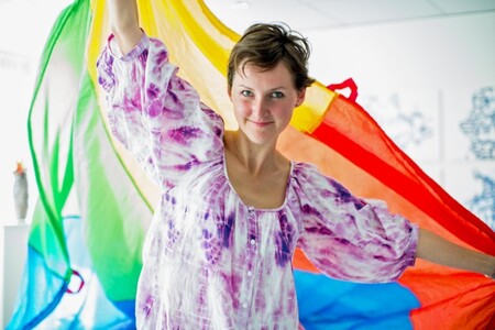 En person med lila och vit tunika håller med utsträckta armar en regnbågsflagga bakom sin rygg.