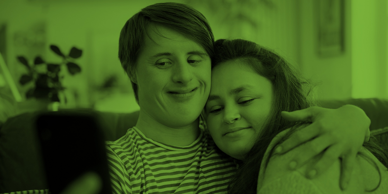 Två personer sitter i en soffa och tar en selfie tillsammans, de kramar om varandra och ser glada ut.