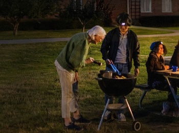 En grupp människor grillar och sitter vid ett bord upplyst av batterilampa utomhus på kvällen framför ett flerbostadshus.