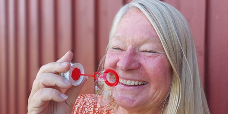 Leende kvinna blåser såpbubblor framför en röd trävägg.