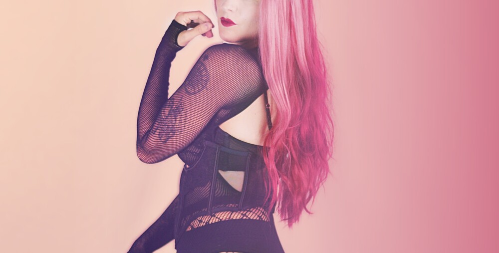 Person med långt rosa hår och svart nätklädsel poserar mot rosa bakgrund.