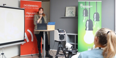 Kvinna håller föredrag med mikrofon framför två rollups, en röd och en grön, i ett ljust rum.