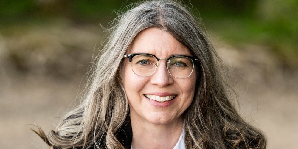 Leende kvinna med långt grått hår och glasögon utomhus, klädd i vit skjorta och mörk kavaj.