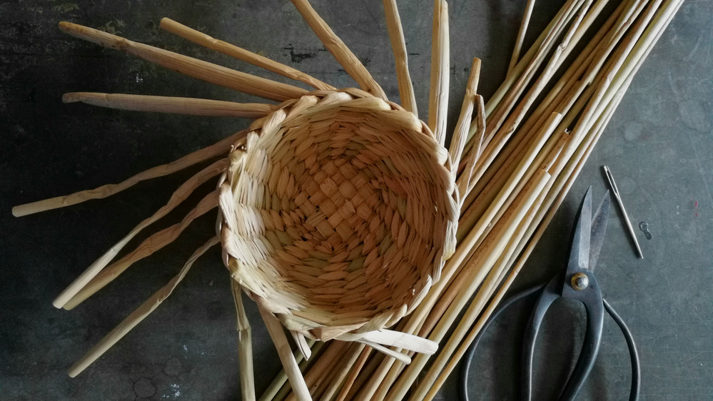 En halvfärdig flätad korg med rottingpinnar, sax och nål på ett bord.