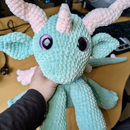 En hand håller ett turkost och rosa stickat gosedjur som ser ut som en fantasidrake med stora ögon.