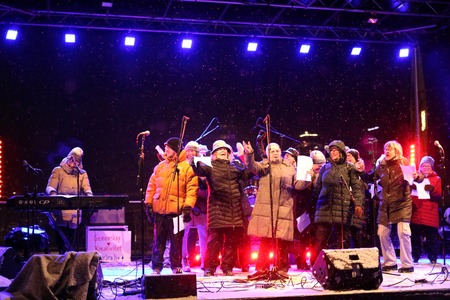 En grupp människor sjunger på en utomhusscen i vinterkläder under kvällstid, med mikrofoner och scenbelysning.