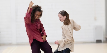 Två personer dansar tillsammans i ett ljust rum med vita väggar och löst sittande kläder.