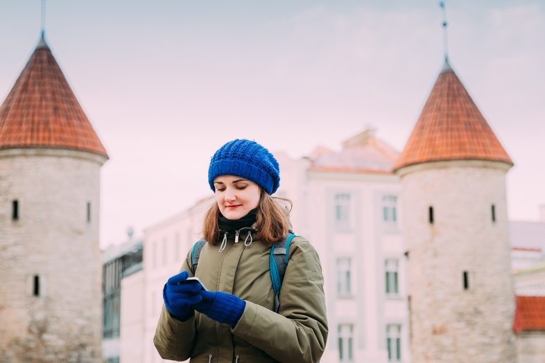 Ung kvinna med blå mössa och vantar står framför medeltida torn och tittar på sin mobiltelefon.