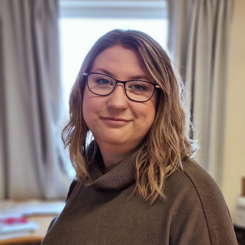 Kvinna med axellångt blont hår och glasögon står inomhus framför ett fönster med gardiner.