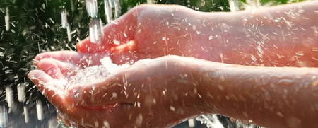 Två kupade händer fångar rinnande vatten utomhus med grönt i bakgrunden.