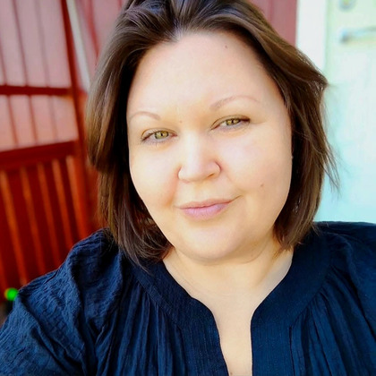 Kvinna med brunt hår och svart blus tar en selfie utomhus framför en röd vägg.
