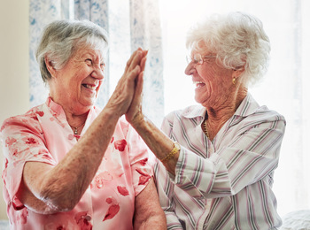 Två äldre kvinnor skrattar och ger varandra en high five i ett ljust rum.