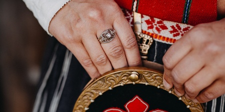 Närbild av händer som håller en traditionell väska med röd brodyr och mässingsdetaljer, klädd i folkdräkt.