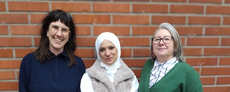 Tre personer står framför en tegelvägg utomhus, klädda i vardagskläder.