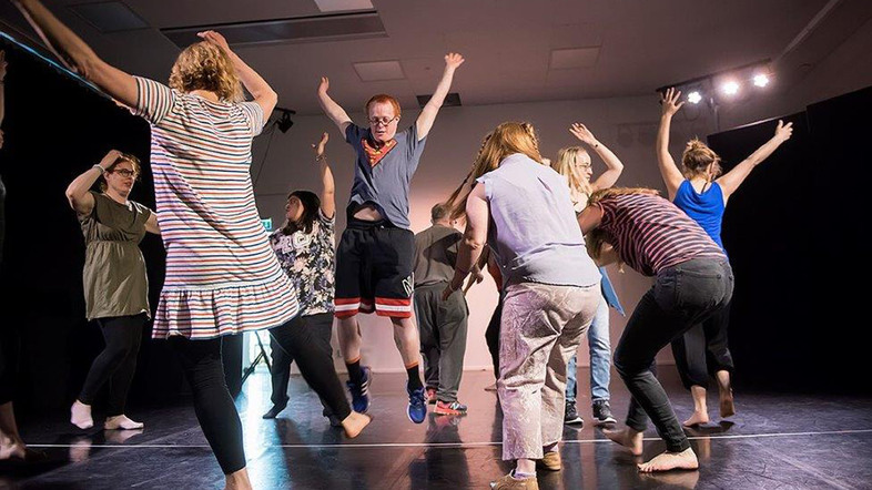 En grupp människor dansar och hoppar tillsammans i en ljus sal med svart golv.