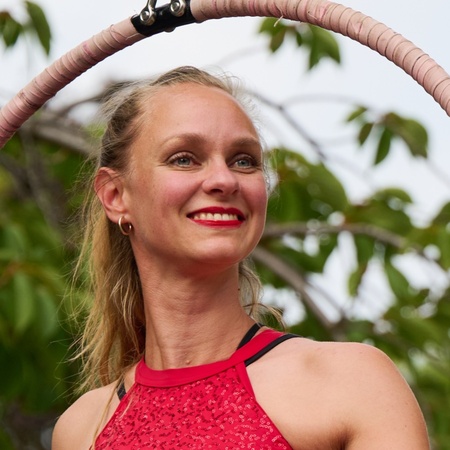 Kvinna i röd topp poserar leende i en cirkusring utomhus med gröna träd i bakgrunden.