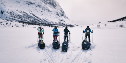 Fyra personer med pulkor står på skidor i ett snötäckt fjällandskap med berg i bakgrunden.
