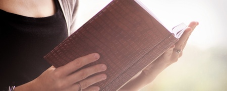 En person håller och läser en bok med brun pärm, endast överkroppen och händerna syns.