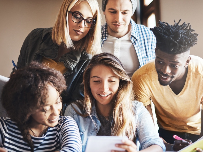 Fem unga vuxna studerar tillsammans vid ett bord, tittar på papper och ler.