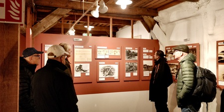 Fem personer tittar på historiska utställningstavlor i ett museum med träbjälkar och rödbruna väggar.