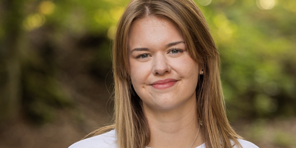 Ung kvinna med ljust hår och vit t-shirt står utomhus med suddig grön bakgrund.