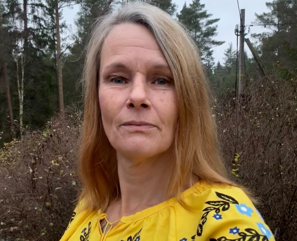 Person med långt blont hår i gul blus med blommönster står utomhus i en skogsmiljö.