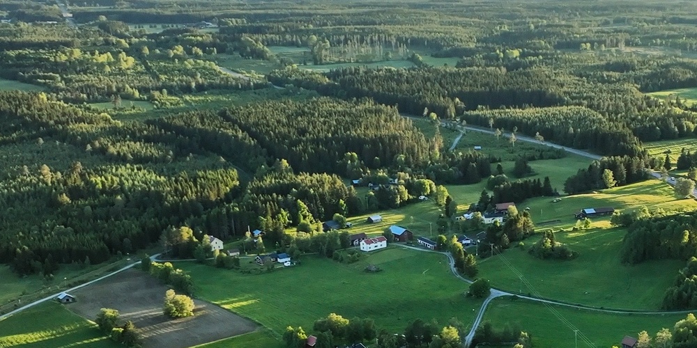 Flygfoto över ett grönt landskap med åkrar, skog och utspridda hus i kvällsljus.