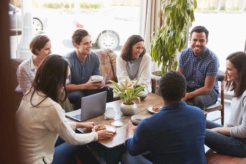 Nio personer sitter i en soffgrupp och fikar tillsammans. På bordet står en laptop och en krukväxt.