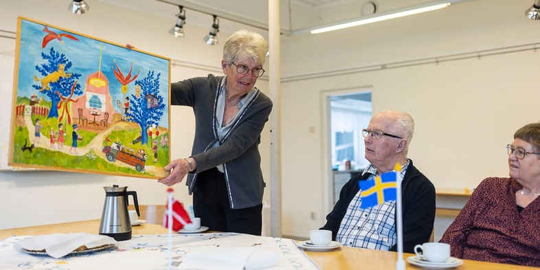 En äldre kvinna visar en färgglad tavla för två äldre personer vid ett bord med kaffekoppar och svenska flaggor.