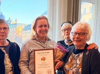 Fyra kvinnor står tillsammans inomhus, en håller ett diplom och blommor, alla ser glada ut.
