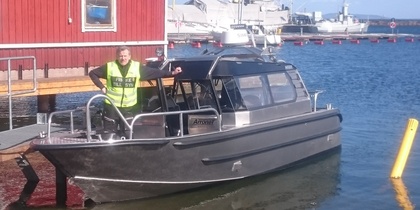 En person i gul väst står på en metallbåt vid en brygga med röda byggnader och båtar i bakgrunden.