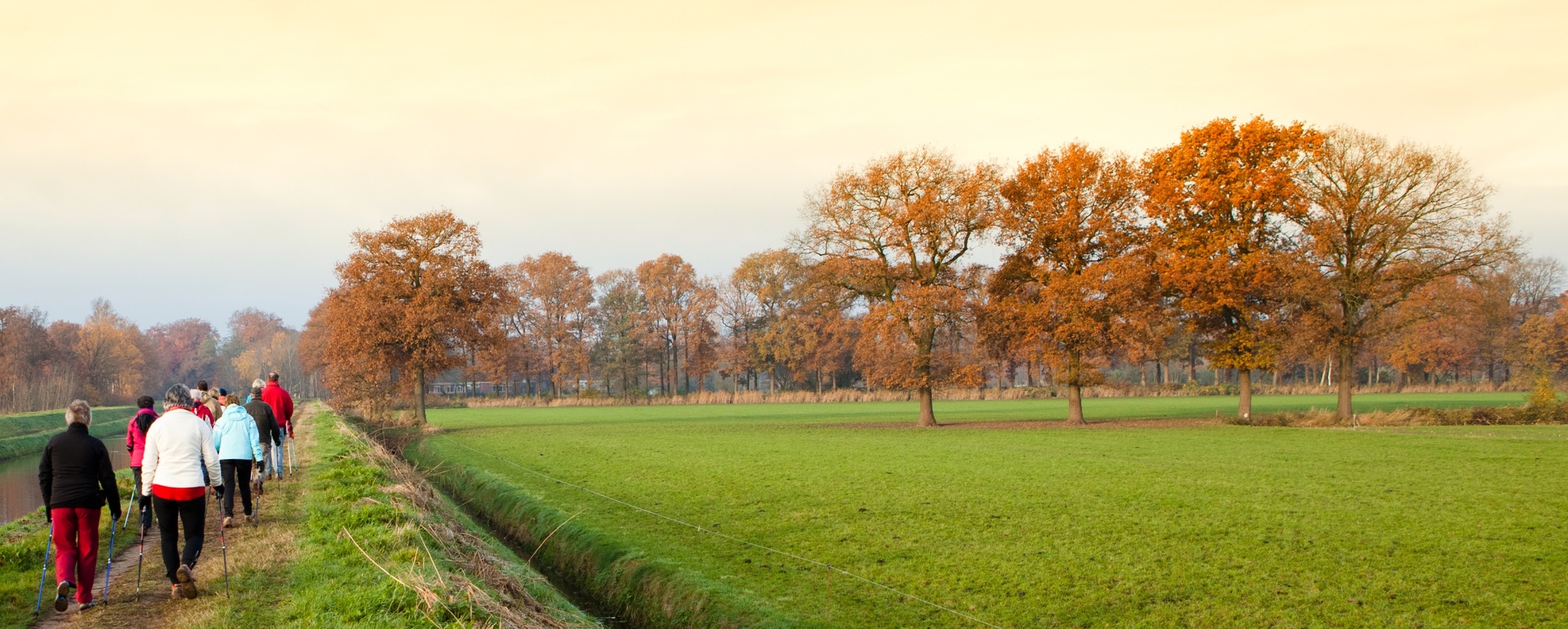 En grupp människor promenerar längs en kanal i ett höstlandskap med gröna fält och träd med orange löv.