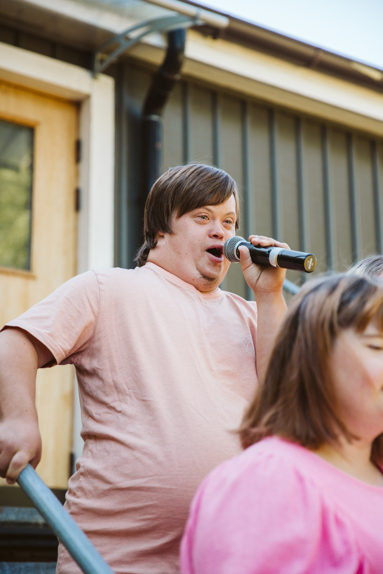 En man i ljusrosa tröja sjunger i en mikrofon utomhus vid ett hus, med en annan person i förgrunden.