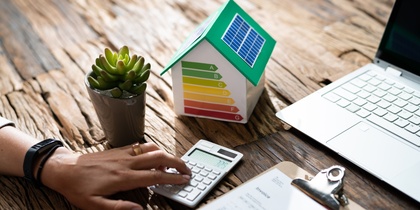 En person använder miniräknare vid ett skrivbord med laptop, pappersarbete, växt och modellhus med energimärkning.