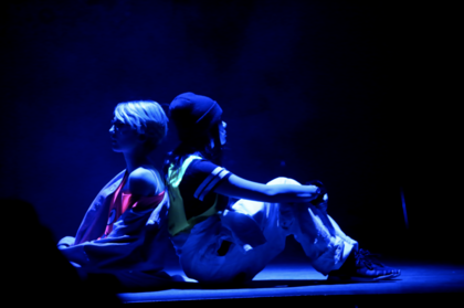 Två personer sitter rygg mot rygg på en scen i blått ljus, klädda i avslappnade kläder.