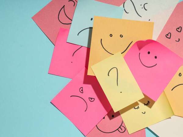 Postit-lappar i olika färger med olika ritade känslouttryck sitter på varandra huller om buller på en turkos bakgrund.