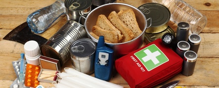Nödutrustning med konserver, vattenflaskor, första hjälpen-kit, ficklampa, kniv, ljus och mediciner på ett träbord.