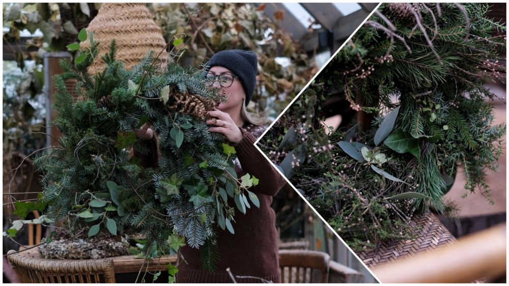 Person håller en stor grön krans av granris och blad i en rustik miljö med naturmaterial.