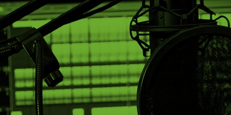 En mikrofon med popfilter framför en datorskärm som visar ljudredigeringsprogram, i grönt ljus.