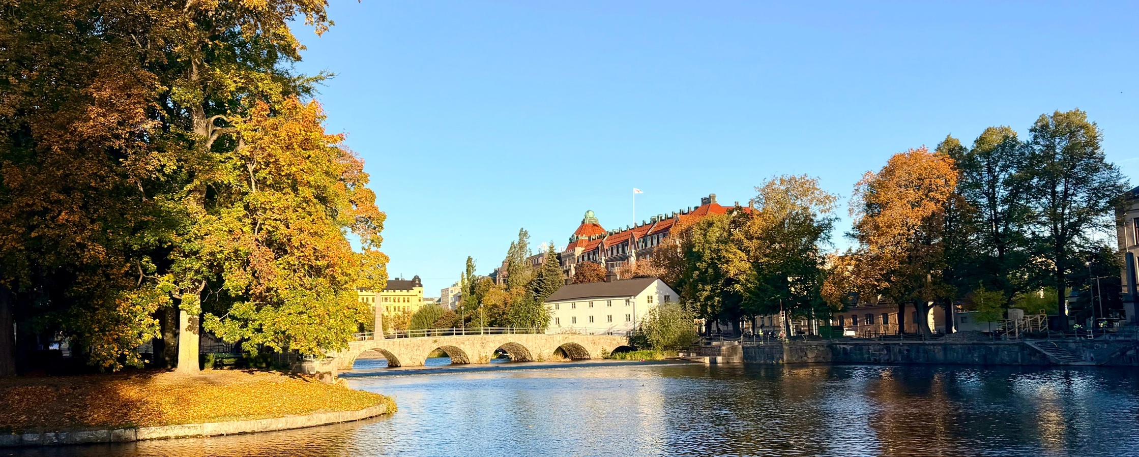 En flod med en stenbro, omgiven av höstfärgade träd och byggnader under en klar blå himmel.