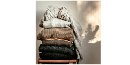 En stapel med vikta stickade tröjor i olika jordnära färger på en stol.