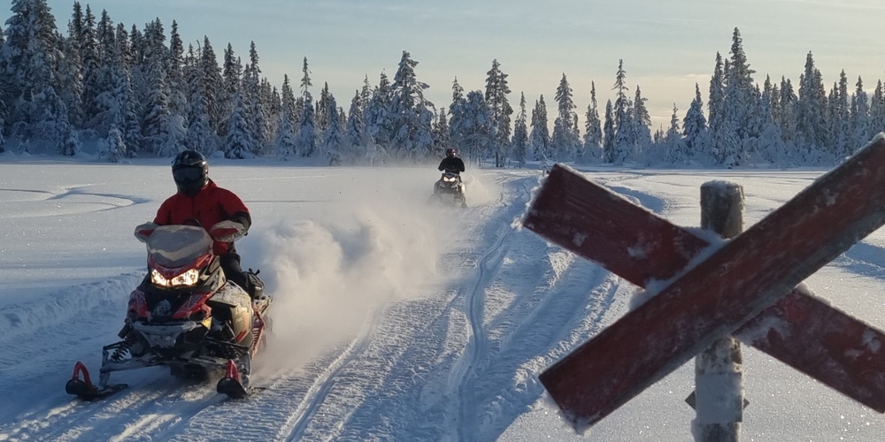 Två personer kör snöskoter på ett snötäckt fält med skog i bakgrunden och en röd krysskylt i förgrunden.
