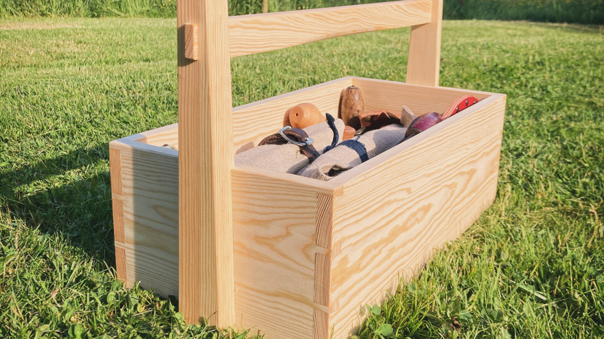 En trälåda med handtag står på gräsmatta, fylld med olika verktyg och föremål.