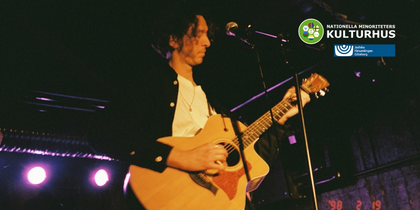 Person spelar akustisk gitarr på scen under en konsert, med logotyper för Nationella Minoriteters Kulturhus och Judiska Församlingen Göteborg.