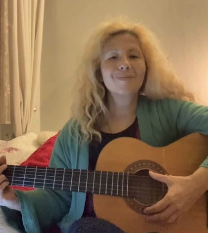 Kvinna med ljust lockigt hår sitter inomhus och spelar akustisk gitarr.